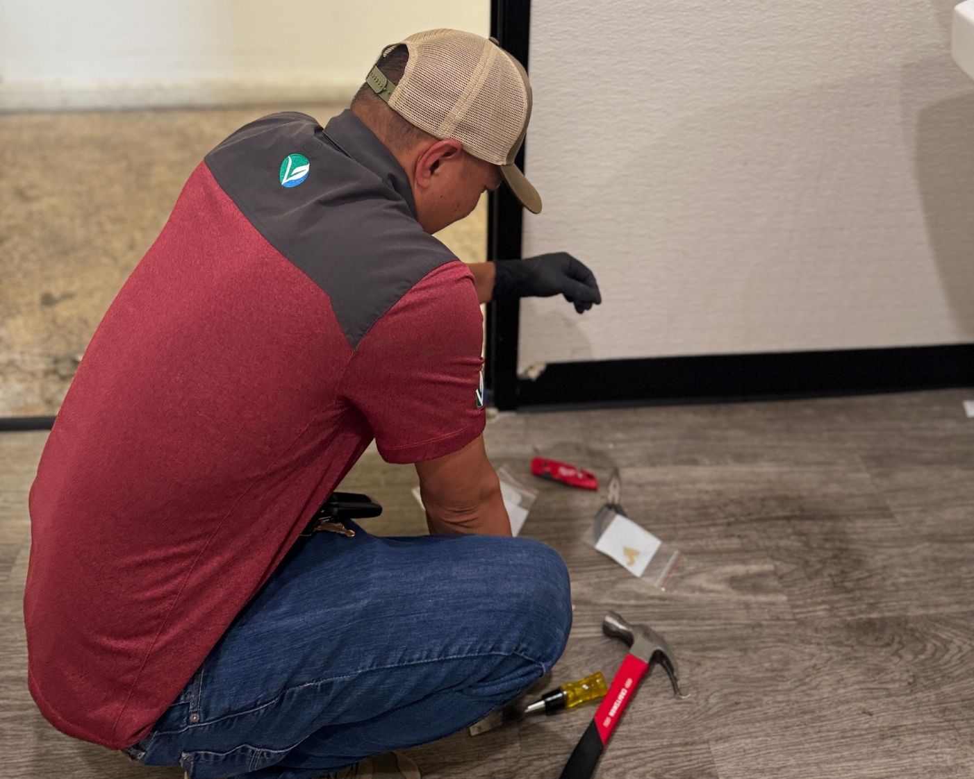 user-inserted Technician performing asbestos field sampling, kneeling on the floor with tools including a hammer and testing equipment, wearing protective gloves and company uniform during an indoor inspection.