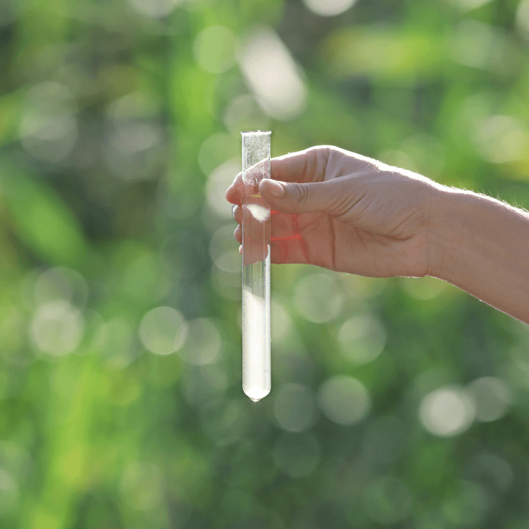 Environmental water sampling and testing: A clear glass vial containing a liquid sample held up against a natural outdoor background.